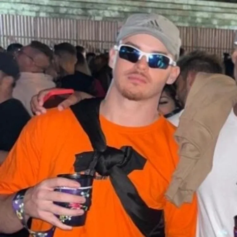 O Professor numa festa de camiseta laranja e óculos espelhados, segurando um copo, com uma mão no ombro dele